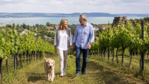 Ehepaar mit Hund spaziert durch die Weinberge am Balaton in Ungarn, mit Blick auf den See und die Hügellandschaft im Hintergrund.